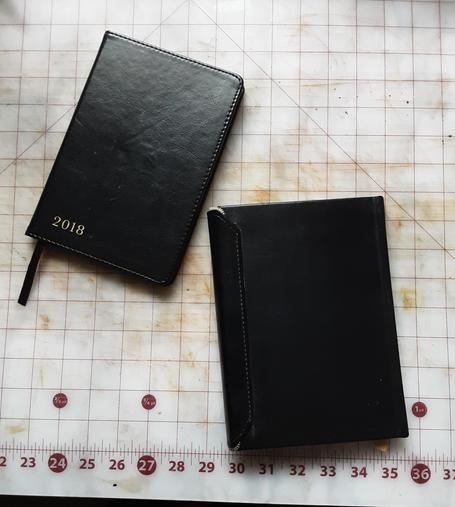 The old date book is in the upper left corner, minus the closure flap. A new black leather book is in the lower right corner with the back facing upward to show the envelope flap stitched to the back left side.