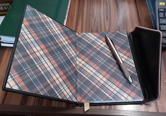The new book lay open to the front cover page, showing the black, navy, red, tan and yellow plaid end papers. The pattern is slanted at a 45 degree angle. The closure flap is open on the right side. There is a muted gold pen laying on the right side of the open book.