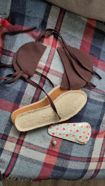 Espadrille shoe soles and brown linen pieces that will be the coverings, all laying on a blue and red checkered blanket. Next to them are an embroidery scissors cover with a floral print and a metal thimble.
