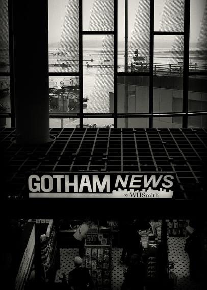 A black and white photo showing an airport terminal window view. In the foreground, a sign reads 'Gotham News' above a store. Past the window, airport ground operations and distant planes are visible. Patterns of light and shadow play across the scene