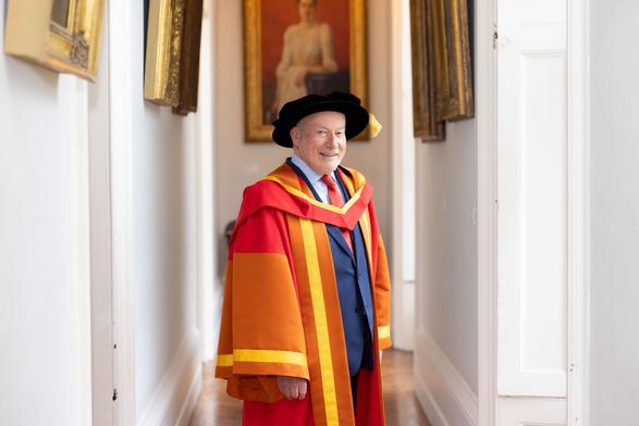 Photograph of a man in a blue suit under gold, orange and red academic robes. He is seen indoors and is wearing a black ceremonial hat with a gold tassle