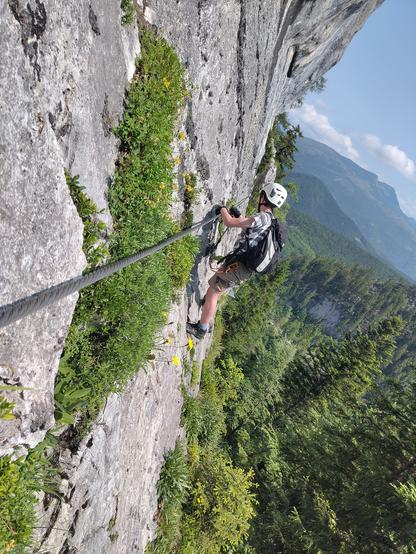 Junger Mensch in Klettermontur steht auf steil abfallendem Fels und hält sich am Stahlseil. Im Hintergrund weitläufiger Ausblick auf andere Berge in der Ferne.