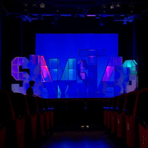 Bühne im Jungen Theater mit dem Aufbau von großen Buchstaben, die das Wort Samstag erheben, alles ist dunkel, der Hintergrund blau mit leichten Kontrasten von Lila und Grün, man sieht die roten Sessel und den Mittelgang bis vorne.