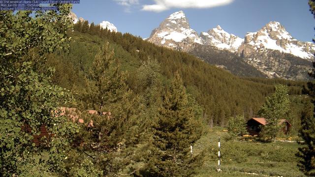 Trees and mountains fill this view from Grand Teton National Park. Tucked away in the trees are two of the cabins found at the Grand Teton Climbers’ Ranch. Snow can be seen in the mountains in the distance, while an alternating black and white snow ruler stands in the vegetation in the foreground. The sky is generally blue with a few white clouds here and there.
