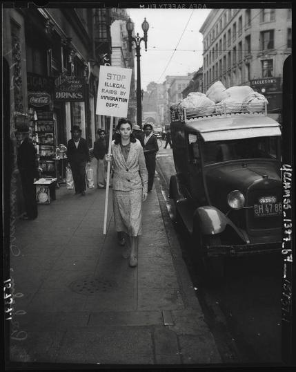 Black and white photo of Los Angeles 1937 showing white woman with sign saying "stop illegal raids by immigration dept." with black men behind looking on.