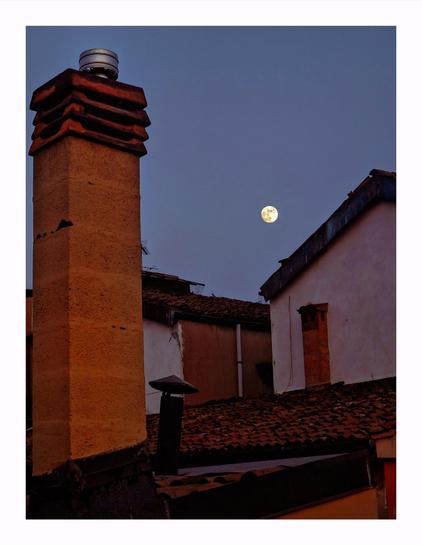 This photo is of a evening scene of rustic rooftops and chimneys, in Bologna. On the left hand side of the photo there is a prominent stone chimney with a metal cap stands in the foreground. In the mid to background there are other terracotta-tiled roofs and stucco walls recede back and fill about half of the photo. Above them, a full moon is present against a deep blue sky. The moon is located on the right hand side of the image just above a diagonally sloping roof.