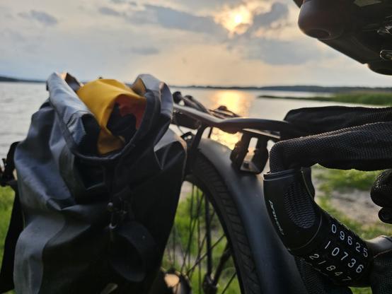 Ein dunkles Fahrrad mit gelber Tasche an einem See. Im Hintergrund geht langsam die Sonne unter. Am Fahrrad ist am Zahlenschloss das heutige Datum in der Form 09625 eingestellt.