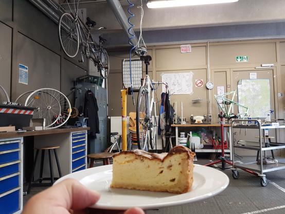 Im Vordergrund ein Stueck Kaesekuchen im Hintergrund ein Fahrrad in einem Montagestaender eingespannt.