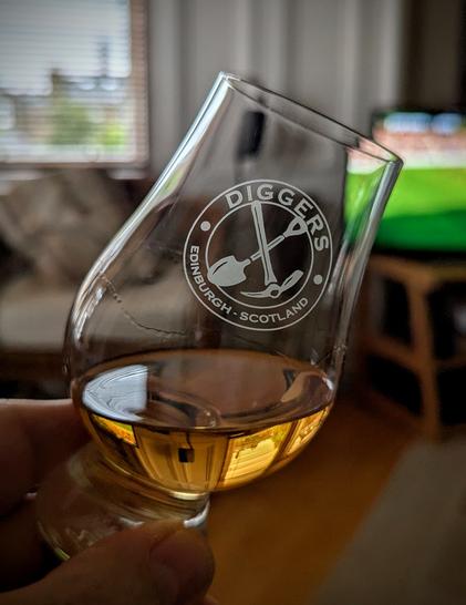 Glencairn glass with a measure of golden coloured whisky. The glass is etched with the words "Diggers, Edinburgh, Scotland" and a depiction of a pick and shovel.