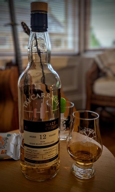 A bottle of Tri Carragh Glen Elgin whisky beside a Glencairn glass on a round table. A blurry image of a guitar in the background.