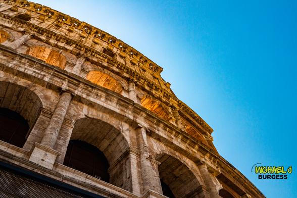 Low-angle view of the Colosseum in Rome during golden hour, with warm sunlight glowing through its arches and a vibrant blue sky above.