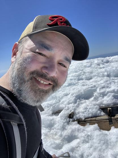 Guy with a short beard and ball cap smiling in front of a pile of snow with some old wood exposed.