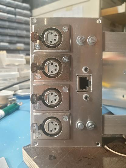 The frontpanel of an electronic device, shown on a workbench with parts drawers in the background. The frontpanel has 4 4-pin xlr connectors and an ethernet connector.