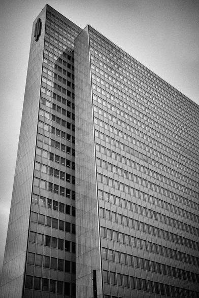 This is a low-angle, black and white photograph of a tall, modern skyscraper. The building dominates the frame, extending from the bottom center upwards towards the top left, creating a sense of immense height. Its facade is composed almost entirely of a grid of numerous square windows, reflecting the light and giving the building a sleek, geometric appearance. The overall impression is one of imposing architecture and modern design.