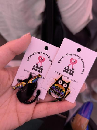 Zwei Pins aus Metall. Eine schwarze Katze mit zwei Regenbogenfischen vor den Augen. Eine schwarze Katze mit buntem Bauch. Darauf steht "sie ihr"