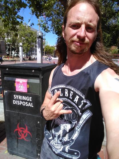 Longhaired guy in a Volbeat tank top featuring a skull with a pompador and razor blades throws horns while posing next to a syringe disposal box with two biohazard symbols on it.