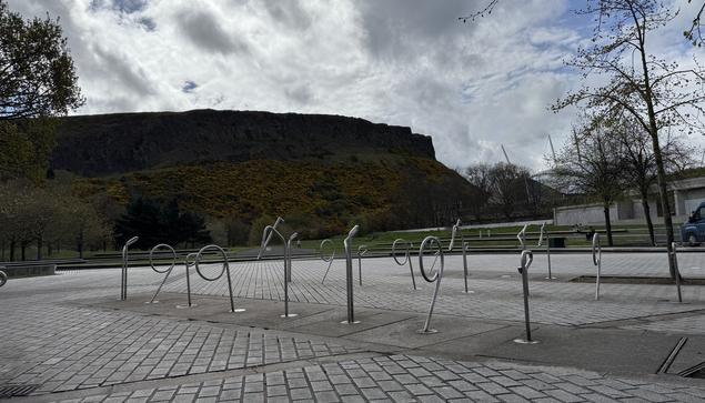 Under a cloudy sky with patches of brilliant blue showing through, Salisbury crags loom over the concrete public space outside the Scottish Parliament, its footing yellowed with gorse bushes in bloom. The stone setts give way to a bank of wildly different shapes bent from metal, creating a small field of unique cycle racks that look more like a public art installation than a functional object. With nothing locked to them, perhaps that’s also not how they’re perceived.