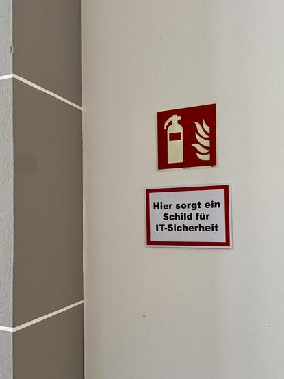 Zwei Schilder an einer hellen Wand in einem Büroflur. Oben ein rotes Piktogramm mit einem Feuerlöscher und Flammen, darunter ein ebenfalls rot umrahmtes Schild mit dem Text: „Hier sorgt ein Schild für IT-Sicherheit“. Die Anordnung wirkt ironisch, da das untere Schild eine Sicherheitswirkung behauptet, ohne konkreten Inhalt zu liefern.