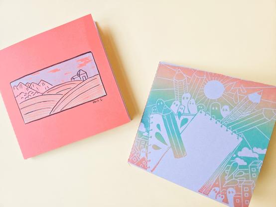 Two small notebooks. On each notebook is a print. One print shows a small landscape, the other shows pencils, notebooks and ghosts.