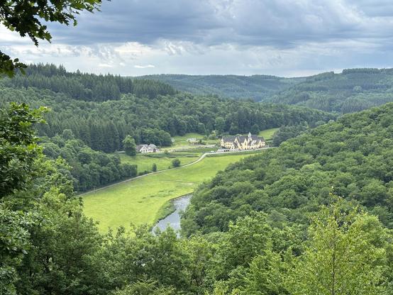 Lushly forested gentle dark green hillsides under a rainy sky surround a grassy light green river valley, in the distance of which a large 19th century beige abbey is visible