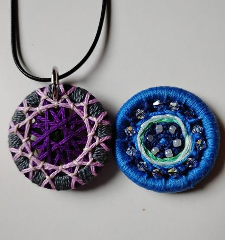 2 thread wrapped buttons lie next to eachother. The one on the left is made of black, dark purple, light purple, grey, with little white details. The one on the right is blue with 12 diamond-like beads along the edge, a stipe of bright green and white, with 6 pearly white beads around the middle of button that has a little dot of lighter blue amongst the dark blue.