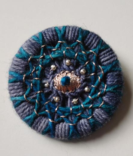 A thread wrapped button made out of a medium blue and turquoise yarn. Every intersection of the strings has a gold stitch over it. 7 golden seed beads are around a golden eye shaped motif in the middle that has a little turquoise French knot on top of it.