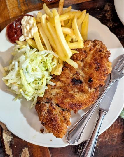 Dunkler Tisch, darauf ein weißer Teller, auf dem sich Salat, Pommes Frites (mit Ketchupt und Mayo), sowie ein großes, goldbraun gebackenes Schnitzel befinden. Besteckt liegt seitlich auf dem Teller.