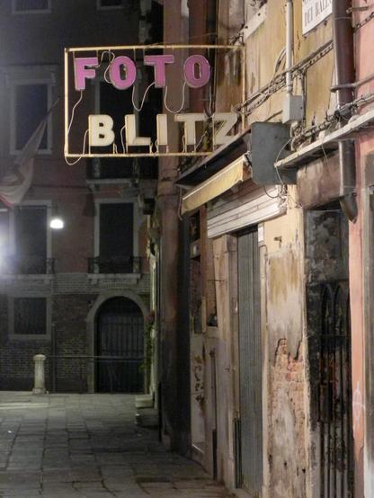 Blick in eine Nebengasse in Venedig bei Nacht. An den Wänden der Gebäude rechts im Bild blättert venedigtypisch der Putz ab. Stromkabel verlaufen auf den Fassaden, Metallgitter verschließen diverse Eingänge. Nichts deutet auf ein Fotogeschäft hin, aber eine nicht leuchtende Neonreklame mit den Buchstaben „Foto Blitz" ragt von den Wänden ins Bild. Sie wirkt genauso in die Jahre gekommen wie ihre Umgebung.
