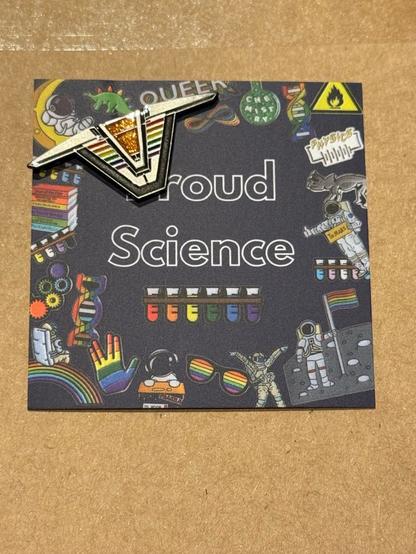 A stargate chevron pin with pride colours on the sides and a glittery orange piece where the chevron would light up while dialing is on a piece of card saying “proud science”