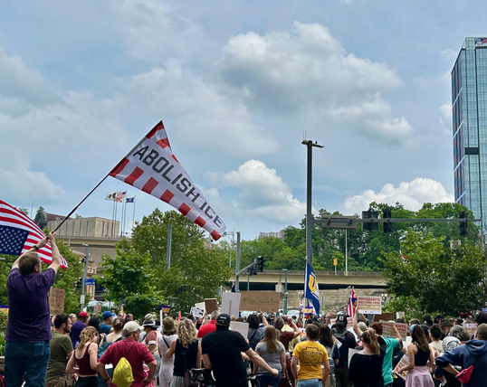 A crowd gathered in downtown Pittsburgh, across the street from the Federal Building. The crowd is carrying signs and flags, including the US flag and one that reads ABOLISH ICE.