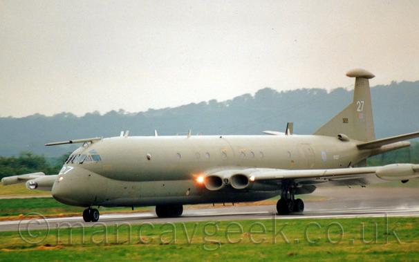 Side view of a 4 jet-engined military patrol aircraft moving at high speed from right to left along a runway, with flaps extended from behind the wings.
The plane has yellowy-grey upper surfaces, and lower surfaces in a slightly lighter shade, and a pale grey belly, with large white numbers "27" on the nose and tail.
There is a small, pastel blue and red roundel on the rear fuselage, next to the white serial "XV227", and a small, pastel red and blue fin flash on the tail.
A bright light is shining in the wing root, on one side of the engine air intakes built in to the wing, with another, smaller, light on the other side of the intakes.
A fixed air-to-air re-fueling probe extends forwards from the top of the cockpit.
Lush green grass fills the foreground, with trees on a hill in the background slowly vanishing in to haze.