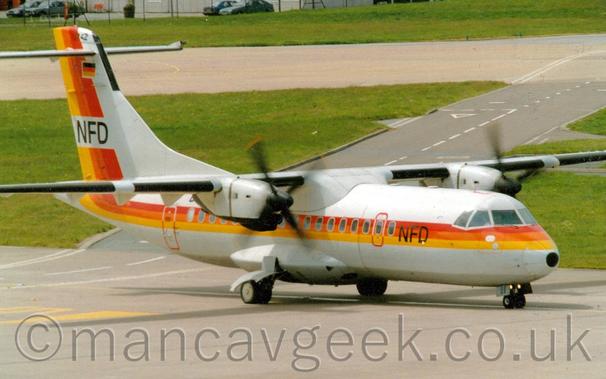 Side view of a high-winged, twin propellor-engined plane taxiing from left to right.
The plane is mostly white, with red, orange, and yellow stripes running along the body, covering the cabin windows, and sweeping up in to the tail, where a white band with letters "NFD" is overlaid.
There are also black "NFD" titles on the forward fuselage.
Grey concrete apron fills the foreground, with grass separating grey taxiways and a black roadway in the background.