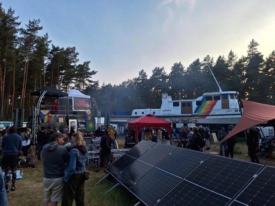 Ein Schiff (die Beluga) steht auf einer Wiese. Links im Bild ist eine kleine Bühne auf einem Auto. Im Vordergrund ist eine Solarpanel und überall dazwischen sind Menschen