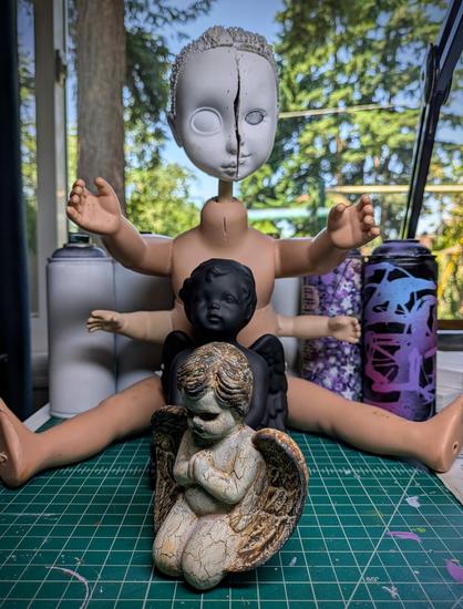 Some works of art in progress with a couple ceramic cherubs getting repainted for a darker style for my Fallen Cherub collection. The item behind the cherubs is a doll with a 2 split heads together and has and extra pair of shorter arms attached just under the doll's normal arms. This work in progress will become a sculpture for my Cyberpunk diety series. And in the background is 6 spray cans primed and ready for illustrations.