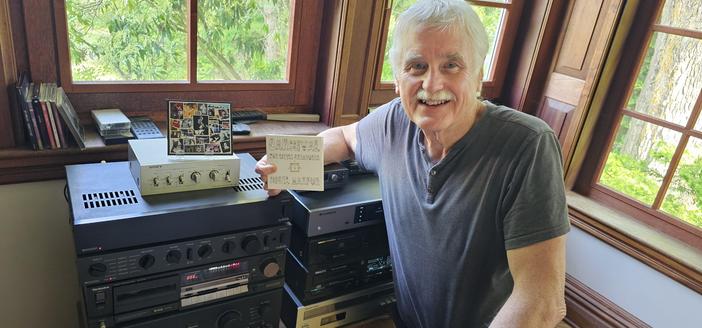 A smiling man with grey hair is in front of stereo equipment in a windowed corner. He is holding a music CD in one hand and there is another case displayed in a holder on top of the equipment.
