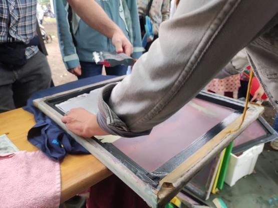 An open air screen printing situation. You can see hands holding a screen, other hands holding a Squeegee with white color on the blade.