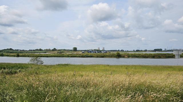 Im Vordergrund eine Wiese mit hohem Gras, dahinter ein breiter Flusslauf. Auf dem gegenüberliegenden Ufer befinden sich Industrieanlagen mit mehreren Gebäuden, Förderbändern und zwei großen Kühltürmen. Darüber ein teils bewölkter Himmel.