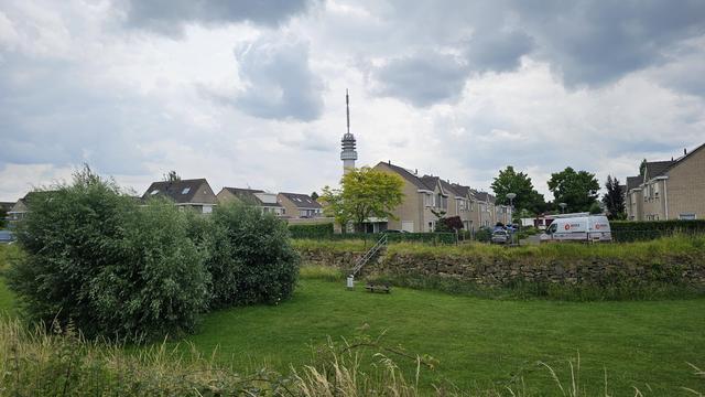 Im Vordergrund eine begrünte Rasenfläche mit niedriger Natursteinmauer, Sitzbank und Metalltreppe. Dahinter eine Siedlung mit Reihenhäusern aus beigefarbenem Klinker. Im Hintergrund ragt ein hoher Sendeturm mit Antenne über die Dächer. Der Himmel ist bewölkt.