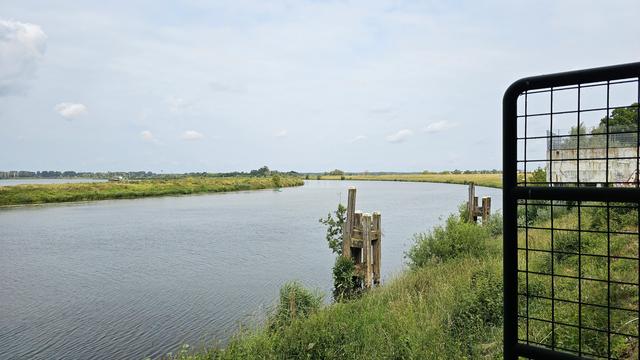 Ein ruhiger Flusslauf mit dichter Ufervegetation, rechts im Vordergrund ein Gittertor und ein verwitterter Betonsockel. Im Wasser stehen alte hölzerne Dalben, dahinter zieht sich das Gewässer in einer leichten Kurve durch die Landschaft.