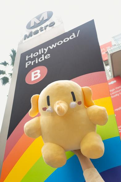Photo of the Creature Mastodon stuffed toy in front of the Hollywood/Pride station sign for LA Metro during a cloudy morning. The Creature is held up in the air by an off-camera arm, its body facing down at the camera. Behind it is the pride flag–decorated Hollywood/Vine station sign, which has been renamed and decorated for LA Pride.