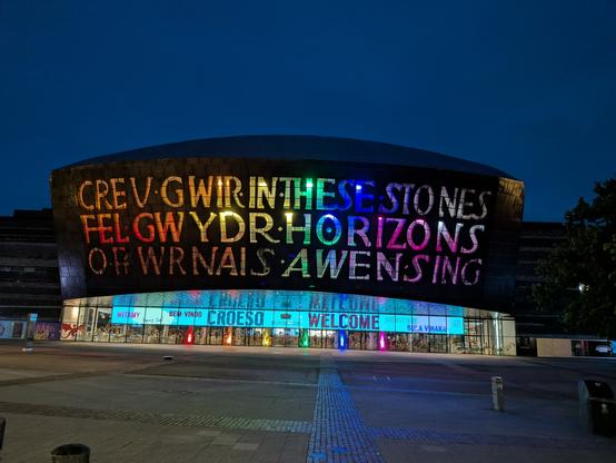 A picture of the Welsh Millennium Centre at night, the sign lit up in Pride colours reading "Creu gwir fel gwydr or wrnais awen," alongside Saesneg, "In these stones horizons sing."