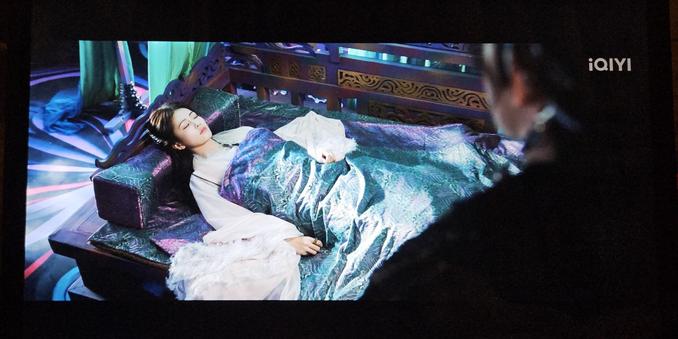 Bai Shuo laying unconscious in a dark purple and turquoise bed with a shiny purple and turquoise duvet that's vaguely iridescent. The floor behind the bed is black with with random streaks of purple and bubblegum pink.