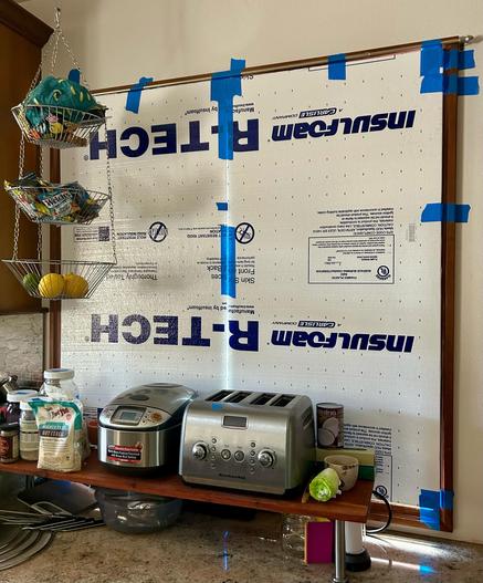 A kitchen scene with an aluminum toaster and rice cooker sitting side-by-side on a shelf with an assortment of hot cereal to the left side of the rice cooker. A three tiered wire basket hangs from the ceiling to the left side and has assorted food & food related items in the baskets. 

In the background a window has been completely covered with two foam insulation panels. The R-Tech panels have been placed with blue tape along the edges. The two panels are also taped together where they meet. 

The panels are 2 feet by 4 feet and 1/2 inch wide. They are printed with blue lettering that has technical data, the brand “insulfoam”, and R-TECH.