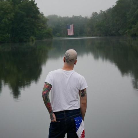 A man with a shaved head stands with his back to the camera. He has a floral tattoo on his head as well as tattoos on his arms. He wears a white t-shirt, jeans, and an American flag bandana in his back right pocket. He gazes out on a lake with a large American flag hanging over it.