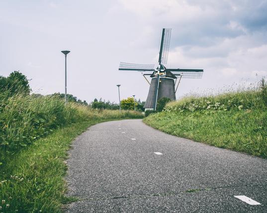 Gray Bicycle path leading to a windmill, blue and cloud sky, with street lamps on the left