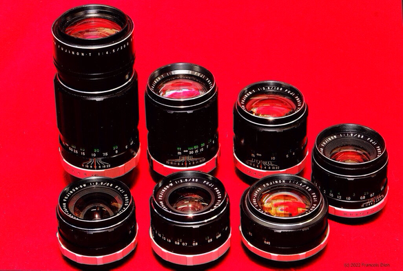 7 lenses arranged in a sideways V with the tallest at the back. 200mm, 135mm, 100mm, 55mm, 50mm, 35mm and 28mm. All feature a common 49mm filter ring. Picture taken with a 55mm fujinon ebc macro lens, manual focus m42