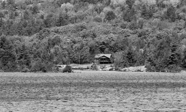 Heavily wooded shoreline across the lake - nestled in an open area, is a home. A flagpole with the Canadian flag stands in an open area. Plane can be seen docked.