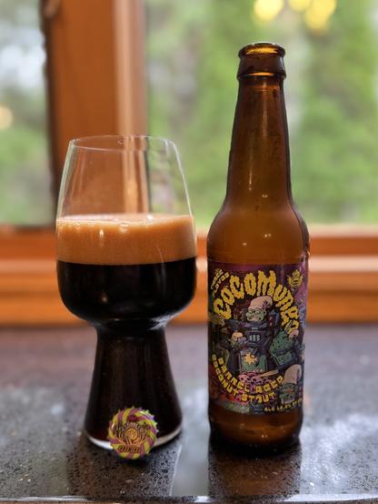 A stout glass filled with of dark beer with a thick dark tan head. The bottle has squiggly yellow text and a Smurf with a machine gun.