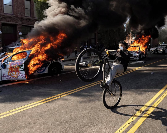 A guy with a medical style face mask does a wheelie on a bicycle along a street. Beside him in the street are three totally trashed and graffitied waymo cars, the two closest are ablaze, you can see huge flames and lots of dark smoke