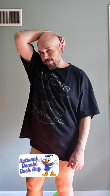 Matt B stands indoors against a gray wall, wearing a black T-shirt with “The Ravens Brewing Co.” printed on it. He has one arm raised behind his head and is gazing at the camera with a slight smile. A “National Donald Duck Day” sticker featuring Donald Duck is placed over his crotch, humorously indicating he’s not wearing pants in honor of the day.